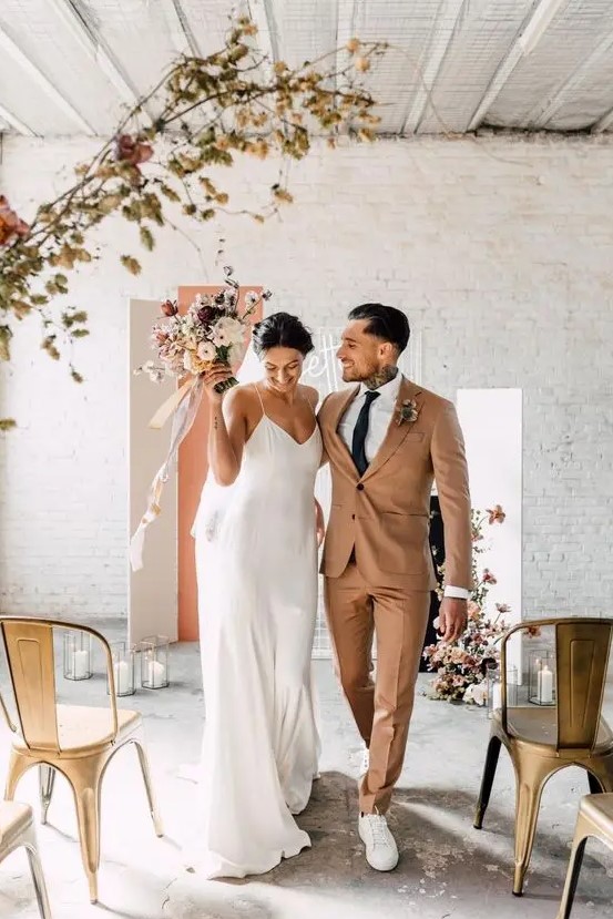 minimalist couple's looks with a rust suit and white sneakers, a white slip maxi wedding dress with a train for the fall