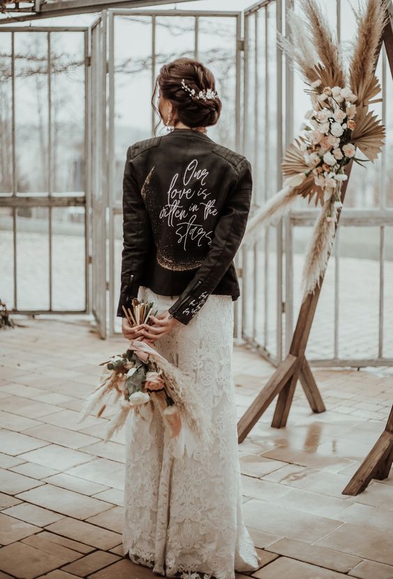 a boho bridal look with a boho lace wedding dress, a black leather jacket with a quote and a boho wedding bouquet with pampas grass