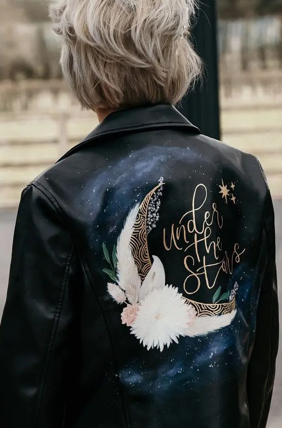 a black leather jacket painted for a celestial wedding, with blooms and a half moon is a gorgeous and romantic cover up idea