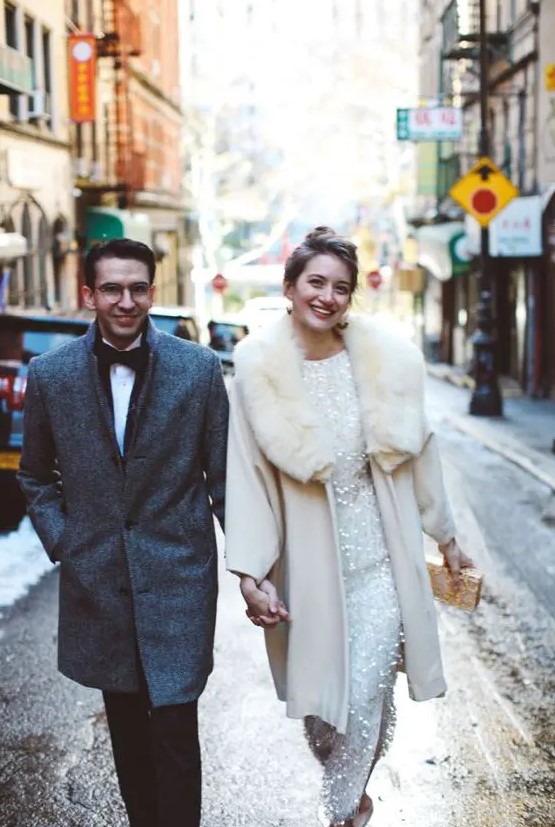 a glam winter bride wearing an embellished wedding dress, a beautiful white coat with a fur collar and rocking a white and gold clutch