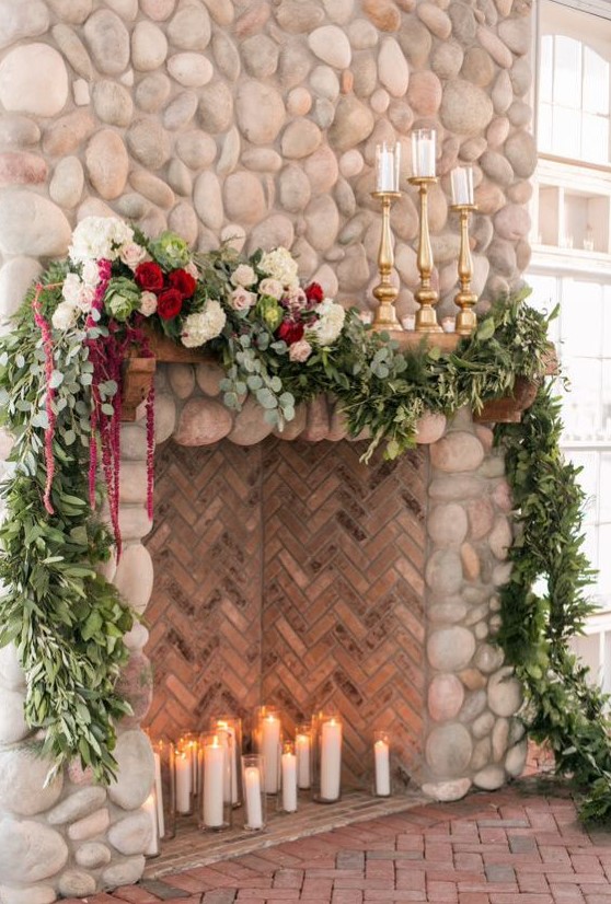 a large fake fireplace deocrated with lush greenery, white and burgundy blooms and with candles on the mantel is a very chic and refined idea
