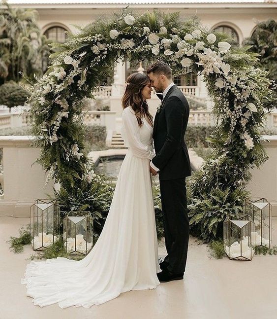 a lovely round wedding arch covered with evergreens and white blooms and candle lanterns around is a cool idea for a winter wedding