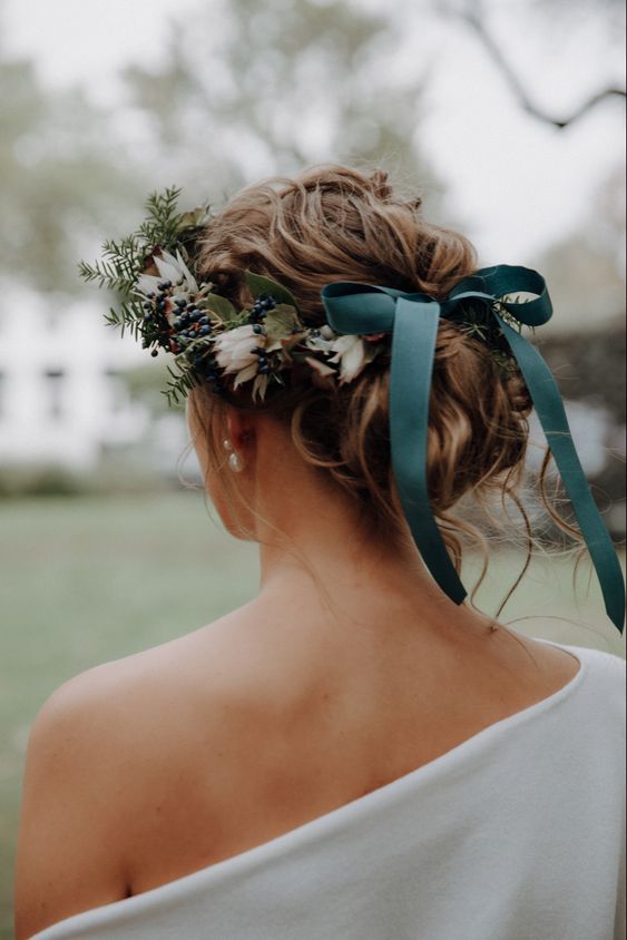 a messy wavy low bun with some locks down, a flower crown with berries and a green bow for a Christmas bride