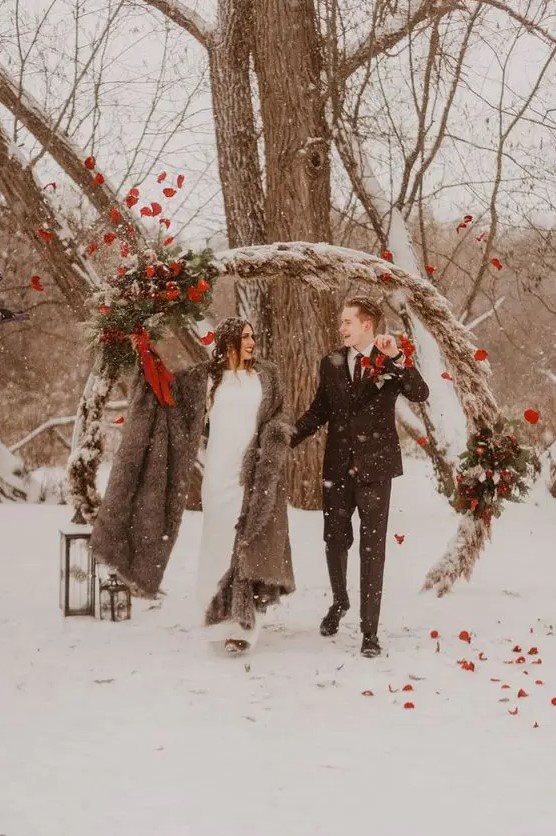 a round pampas grass wedding arch with greenery, bright blooms and red ribbons and pinecones for a winter woodland wedding