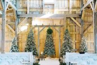 a white barn wedding ceremony space with several Christmas trees with lights and decor, white chairs and evergreens to line up the aisle