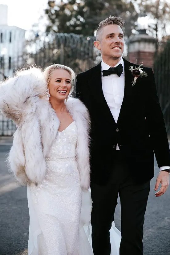 a white embellished lace wedding dress paired with a neutral faux fur coat and statement earrings for a glam and luxurious look