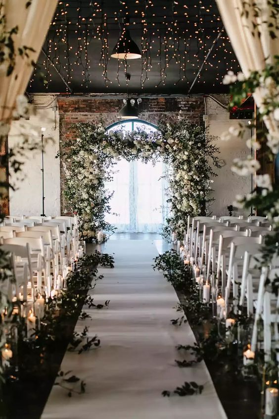 a white winter wonderland wedding ceremony space with a white wedding arch, greenery along the aisle, candles and lights