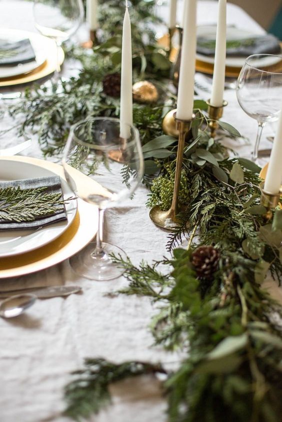 a beautiful and lush evergreen and greenery table runner with pinecones and gold ornamnets plus tall and thin candles is amazing
