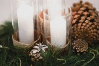 a chic and simple winter wedding centerpiece of evergreens, pinecones, pillar candles in glasses wrapped with bark