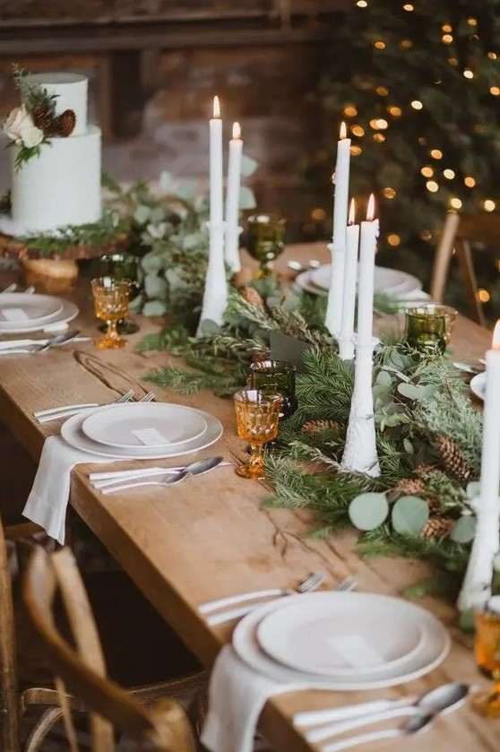 a gorgeous chalet wedding table setting with an evergreen and pinecone table runner, tall candles, white porcelain and amber glasses