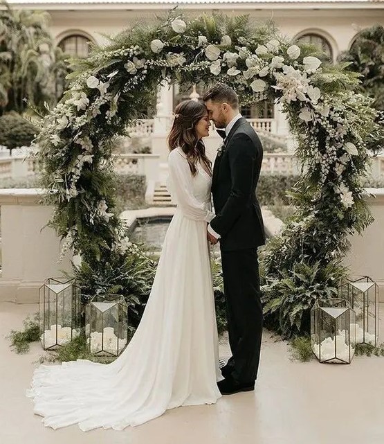 a lovely round wedding arch covered with evergreens and white blooms and candle lanterns around is a cool idea for a winter wedding