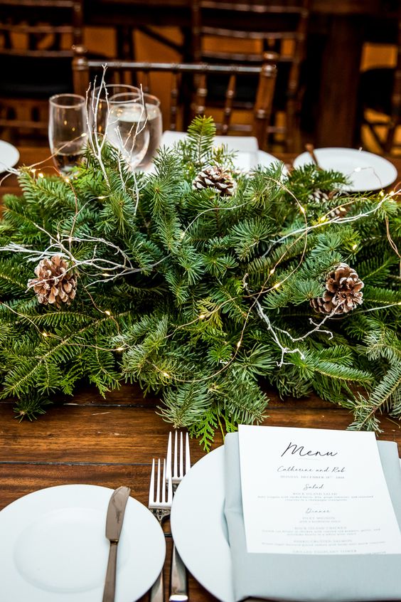 a lush evergreen table runner with snowy pinecones and lights is a super cool and chic idea for a wedding tablescape