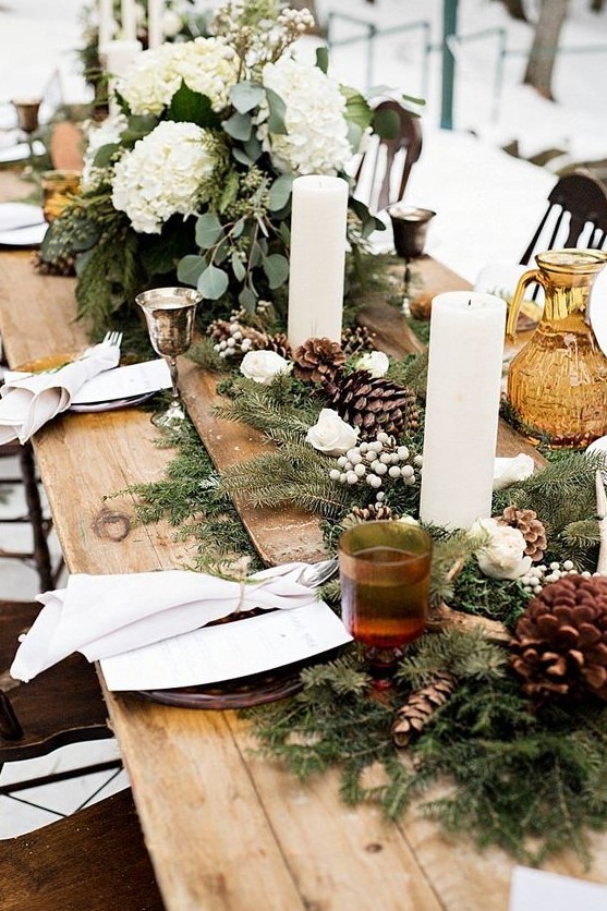 a rustic winter wedding table with an evergreen and pinecone runner, lush white blooms, pillar candles and an uncovered table