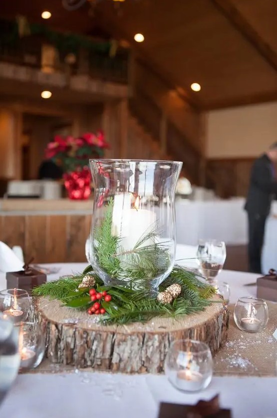 a simple rustic Christmas centerpiece of a wood slice, evergreens, berries, pinecones and candle lanterns