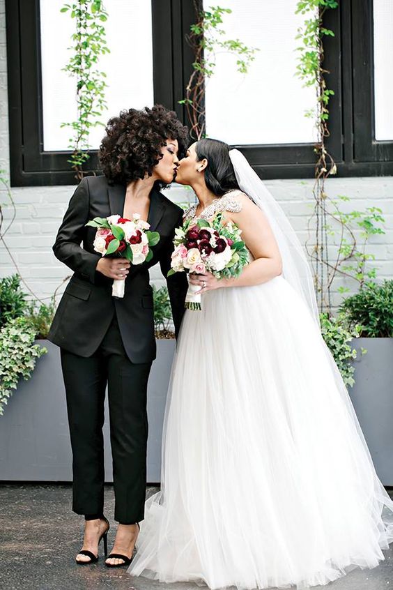 one bride rocking a sexy black pantsuit and heels, the second bride wearing a wedding ballgown with an embellished bodice and a veil