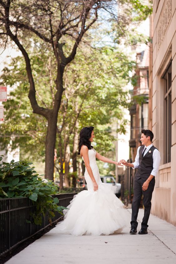 one bride rocking a glam mermaid wedding dress, the second bride rocking grey pants and a waistcoat, a white shirt and a black tie