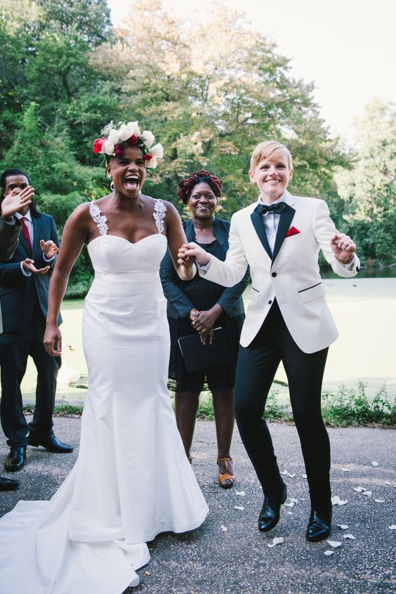 one bride wearing black pants, a white and black jacket, a bow tie, the second bride wearing a mermaid wedding dress with lace straps
