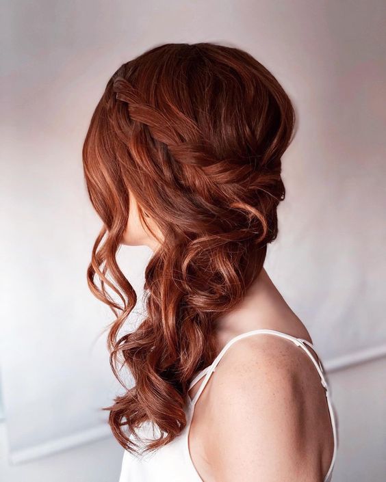 a classy and chic ginger side swept hairstyle with a braided halo, wavy hair down and some waves framing the face
