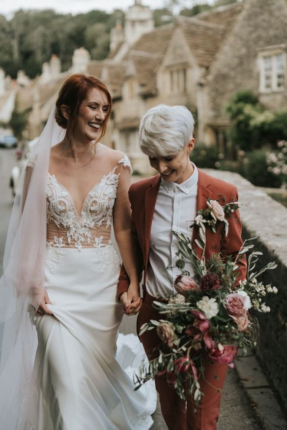 one bride wearing a rust pantsuit with a white shirt, the second bride rocking a wedding dress with an applique bodice and a plain skirt