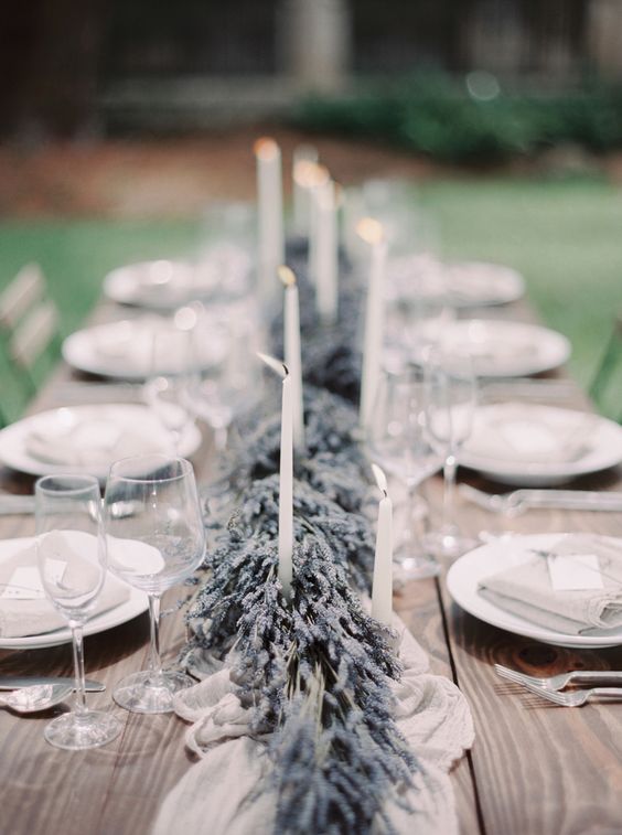 uncovered wooden farm table with a centerpiece of lavender garland, a silk table runner, tall taper candles and place settings of white china