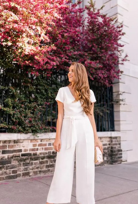 a casual white jumpsuit with a V-neckline, a sash, short sleeves and a neutral bag for a spring or summer bridal shower