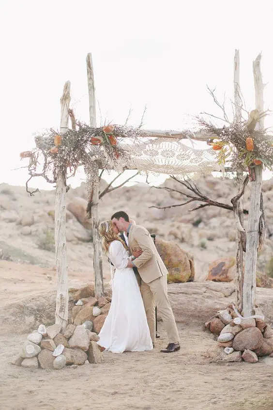 whitewashed wooden chuppah with a macrame hanging above, feathers and flowers