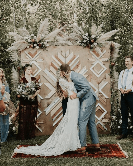 a boho backyard wedding backdrop of plywood, with geometric decorative patterns and some white florals and pampas grass on top