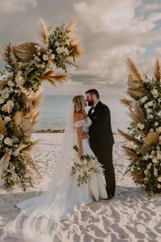 a boho beach wedding altar done with white blooms, pampas grass, fronds and greenery is a cool idea for a beach wedding