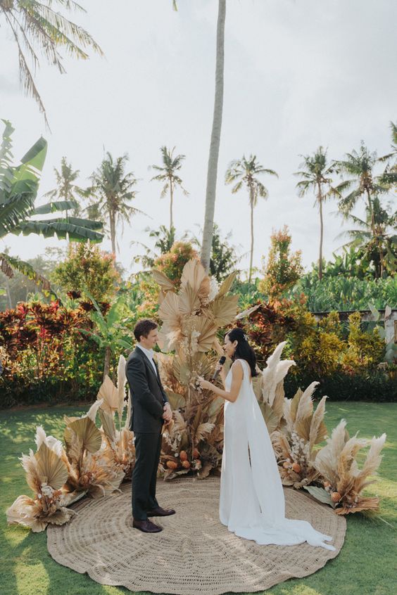 a boho tropical wedding altar with fronds, pampas grass, pink blooms and a jute rug is a cool idea