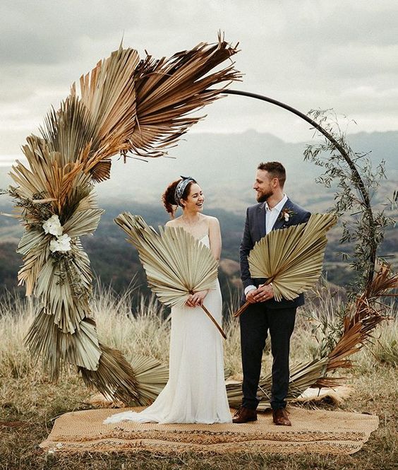 a boho tropical wedding arch covered with fronds and some white blooms is a cool idea for a boho wedding