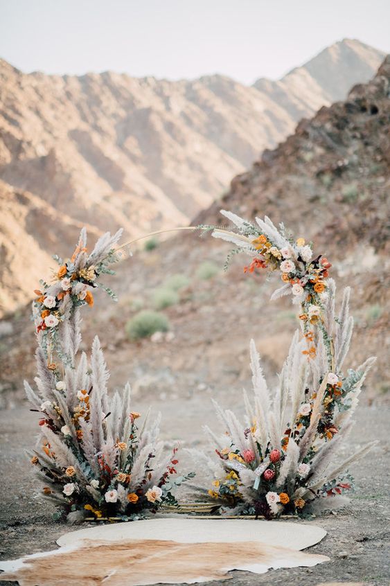 a boho wedding arch with pampas grass, blush, orange and red roses and some king proteas is a cool idea for a boho ceremony