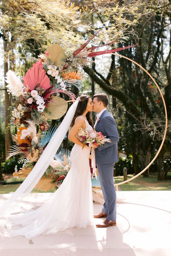 a bright boho wedding arch or a round shape, covered only partly with colorful fronds, blush blooms, colorful grasses