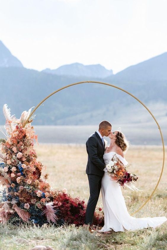 a colorful round wedding arch with bold grasses, blue and pink ones, blush and mauve roses is amazing