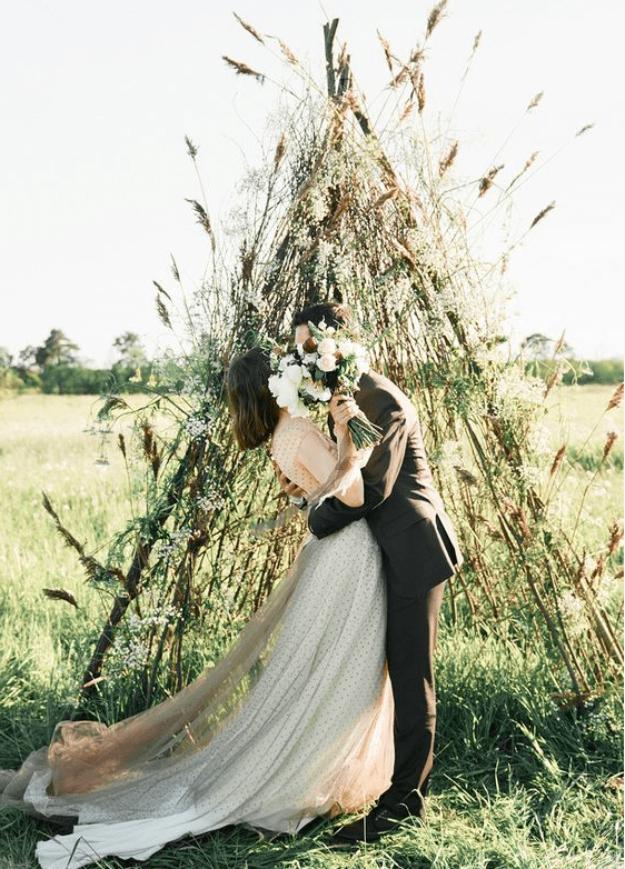 a creative teepee wedding altar made of branches covered with greenery and some dried grasses for a fall boho wedding