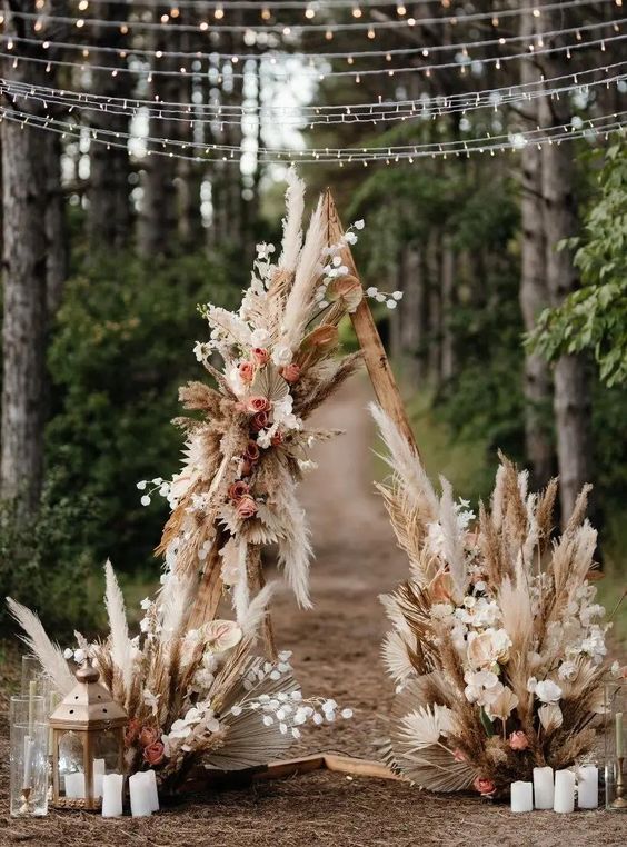 a fantastic triangle boho wedding arch covered with fronds, pampas grass and pink blooms and seed pods and candles is amazing