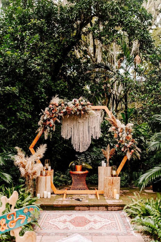 a hexagon boho wedding arch with macrame, yellow, coral and red blooms, pampas grass and greenery plus candles around