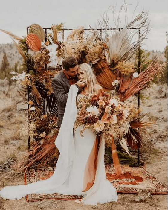 A jaw dropping boho fall wedding backdrop of dried colorful fronds, leaves, bold blooms of mustard, yellow and some white touches