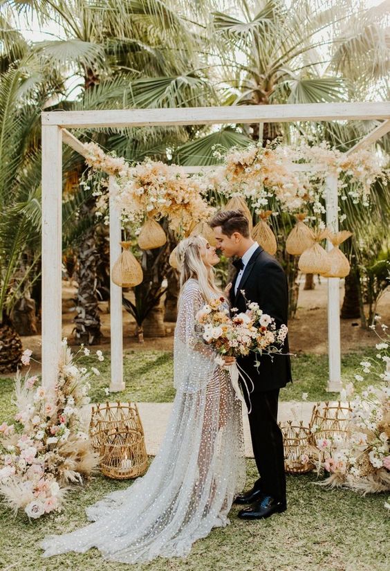 a neutral boho wedding arch with dried blush and neutral blooms, some bags hanging down, lush floral arrangements and candles on the ground