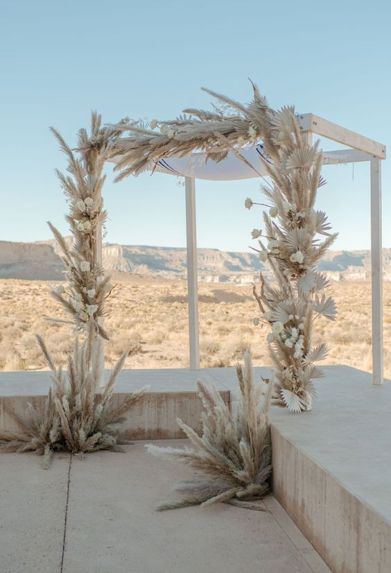 a pure white boho wedding arch covered with pampas grass, white roses and roses is a cool idea for a boho wedding