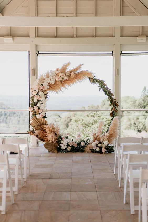 a round boho wedding arch covered with leaves, pampas grass, fronds and white and blush roses and orchids for a refined boho wedding