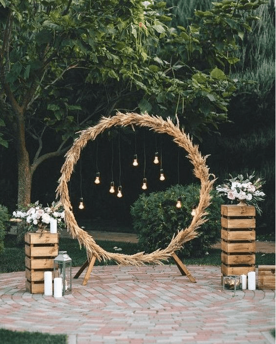a round wedding arch fully covered with pampas grass and with bulbs hanging inside plus blooms and candles on crates