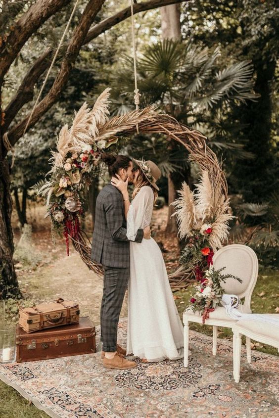 a small round boho wedding arch done with pink and red blooms, pampas grass and greenery is a cool and bold idea for a boho wedding