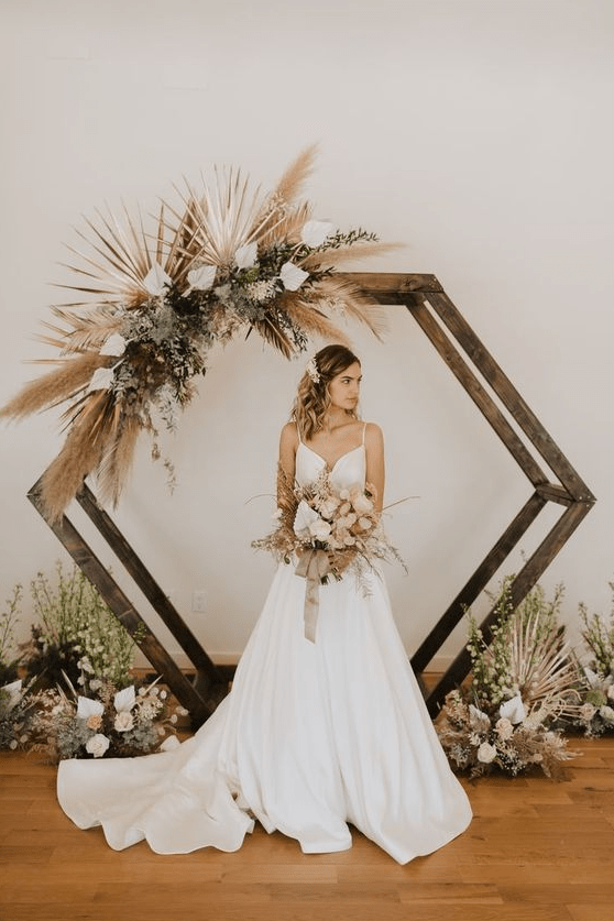 a stained hexagon wedding arch decorated with pampas grass, greenery, white blooms and fronds for a boho wedding