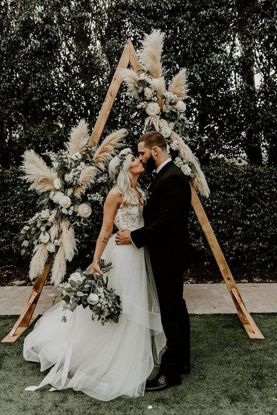 a triangle boho wedding arch with white blooms, greenery and pampas grass is a cool idea for a neutral boho wedding