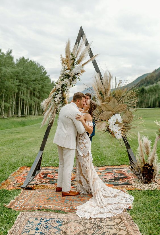 a triangle wedding arch covered with pampas grass, fronds and white blooms plus boho rugs is a great idaa for a summer or fall boho wedding