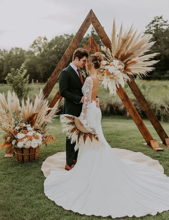 a triangle wedding arch decorated with pampas grass, bold leaves and white and orange blooms is ideal for a flal boho wedding