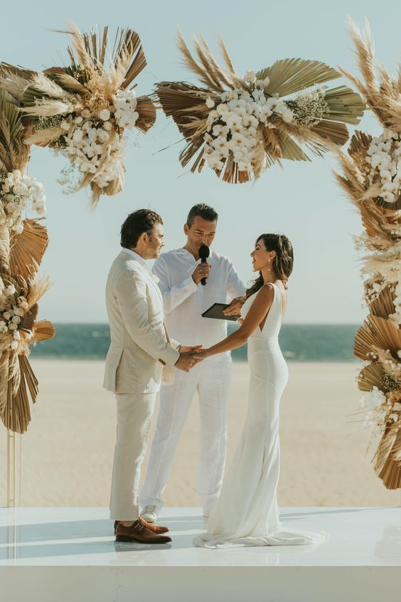 a white boho wedding altar with white orchids, fronds and pampas grass plus an ocean view for a beach boho wedding