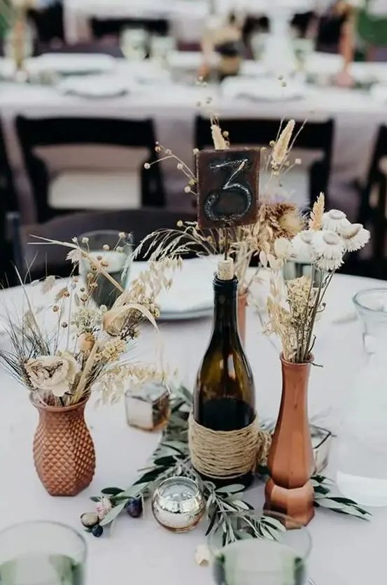 A beautiful rustic wedding centerpiece of rust colored vases, dried blooms, bunny tails, berries, a candle and olive branches is amazing