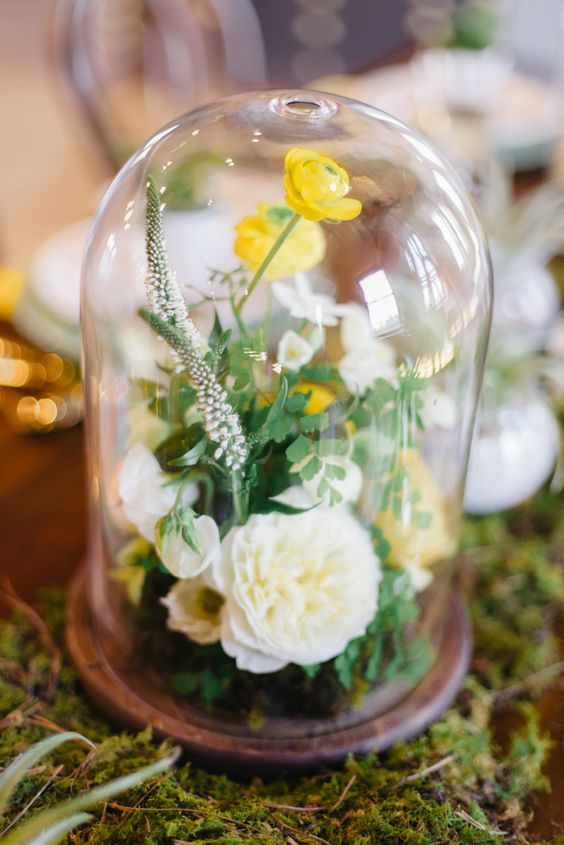 a bright wedding centerpiece of a cloche with greenery, astilbe, white and yellow blooms