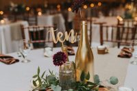 a casual rustic wedding centerpiece of a wood slice, candles, greenery, gilded bottles and burgundy blooms for a backyard celebration